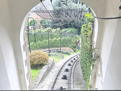 Blick durch das Rundbogenfenster einer Standseilbahn auf die geschwungenen Gleise, umgeben von Grünpflanzen und historischen Laternen.