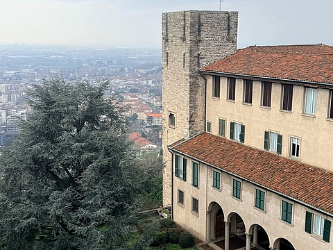 Blick von oben auf einen historischen Kreuzgang mit Arkadengang, Innenhof und mittelalterlichem Turm in der Città Alta Bergamos, dahinter der Blick über die Unterstadt.