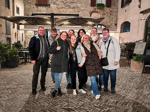 Neun Personen stehen eng zusammen und lachen in die Kamera auf einem nächtlich beleuchteten Kopfsteinpflasterplatz in der Città Alta Bergamos, im Hintergrund ein Restaurant mit Sonnenschirmen.