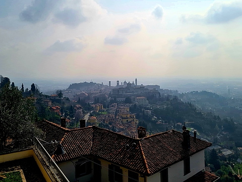 Weitläufiges Panorama über die Hügel und Dächer Bergamos mit der Silhouette der Città Alta und ihren Kirchtürmen im Morgendunst, aufgenommen von einer erhöhten Position.