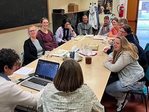Gruppe von etwa elf Personen sitzt an einem langen Tisch in einem Seminarraum mit Tafel und Laptop und führt ein gemeinsames Fachgespräch; die meisten schauen lächelnd in die Kamera.
