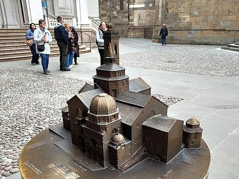 Bronzenes Stadtmodell der Kathedralenanlage Bergamos auf einem runden Sockel auf dem Domplatz, im Hintergrund Besucherinnen und Besucher sowie historische Gebäudefassaden.