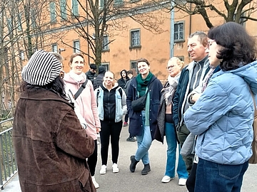 Gruppe von etwa neun Personen steht im Freien auf einem Platz in Bergamo und hört einer Person zu, die von links spricht; im Hintergrund ein historisches ockerfarbenenes Gebäude und kahle Bäume.