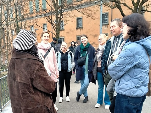 Gruppe von etwa neun Personen steht im Freien auf einem Platz in Bergamo und hört einer Person zu, die von links spricht; im Hintergrund ein historisches ockerfarbenenes Gebäude und kahle Bäume.